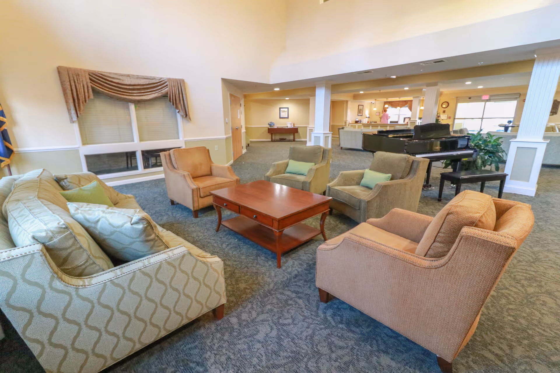 Spacious senior living lounge with sofas and armchairs arranged around a central coffee table and a piano in the background.