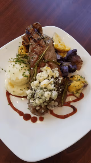 A plated meal featuring a grilled steak topped with crumbled blue cheese and garnished with rosemary, served with mashed potatoes sprinkled with chives, and a side of roasted purple and yellow cauliflower on a white plate with a decorative sauce drizzle.