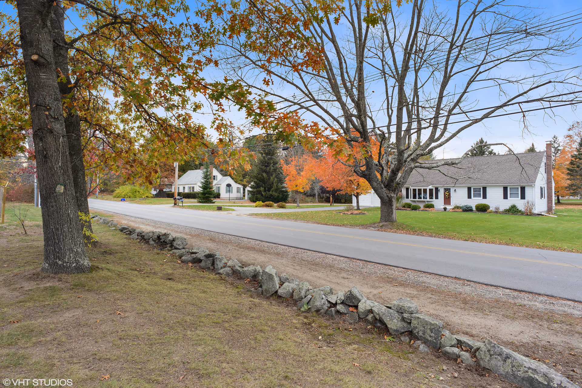 A quiet suburban street lined with trees showing autumn foliage. On the right side of the road, there is a white single-story house with a well-maintained lawn. The sky is clear with a few clouds, and a stone border runs along the edge of the grass near the road.