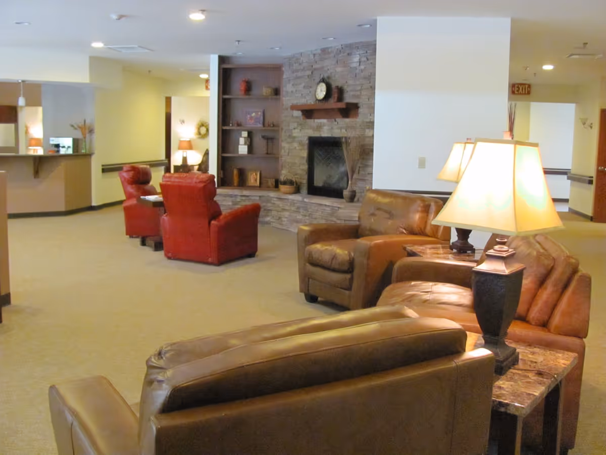 A cozy senior living facility common area with brown leather sofas and armchairs, two red armchairs near a stone fireplace with a clock on the mantel, warm lighting from table lamps, and built-in shelves with decorative items.