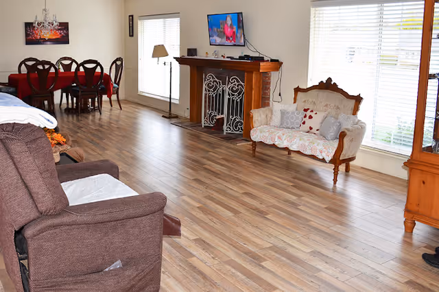 A spacious living room with wooden flooring featuring a brown recliner chair, a vintage-style loveseat with decorative pillows, a wooden fireplace with a TV mounted above it, a floor lamp, and a dining area with a table covered with a red tablecloth and six chairs. Large windows with blinds allow natural light to fill the room.