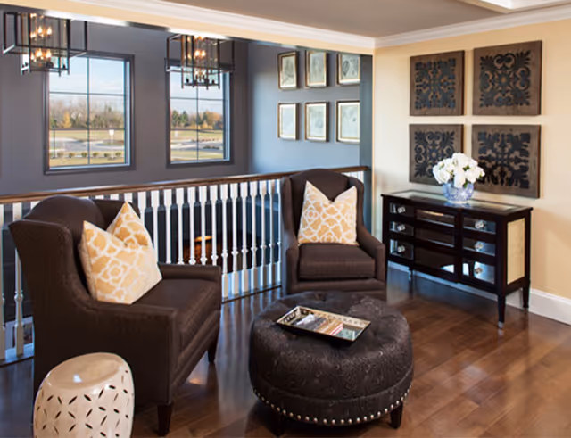 Cozy sitting area with two upholstered armchairs, patterned throw pillows, a round tufted ottoman, and a console table beneath decorative wall art.