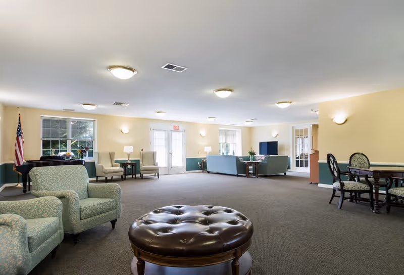 A spacious senior living facility common area with multiple seating arrangements including armchairs, sofas, and a round leather ottoman in the center. The room has beige walls with green wainscoting, several windows letting in natural light, ceiling lights, an American flag near a piano, and a TV on a stand. There are also tables with chairs along the right side of the room.
