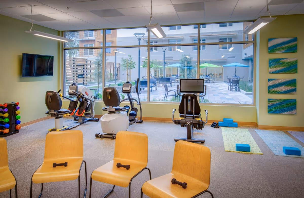 A bright fitness room with exercise equipment including stationary bikes and rowing machines, wooden chairs with small dumbbells on the seats, and yoga mats with blocks on the floor. Large windows show an outdoor patio area with tables, chairs, and colorful umbrellas.