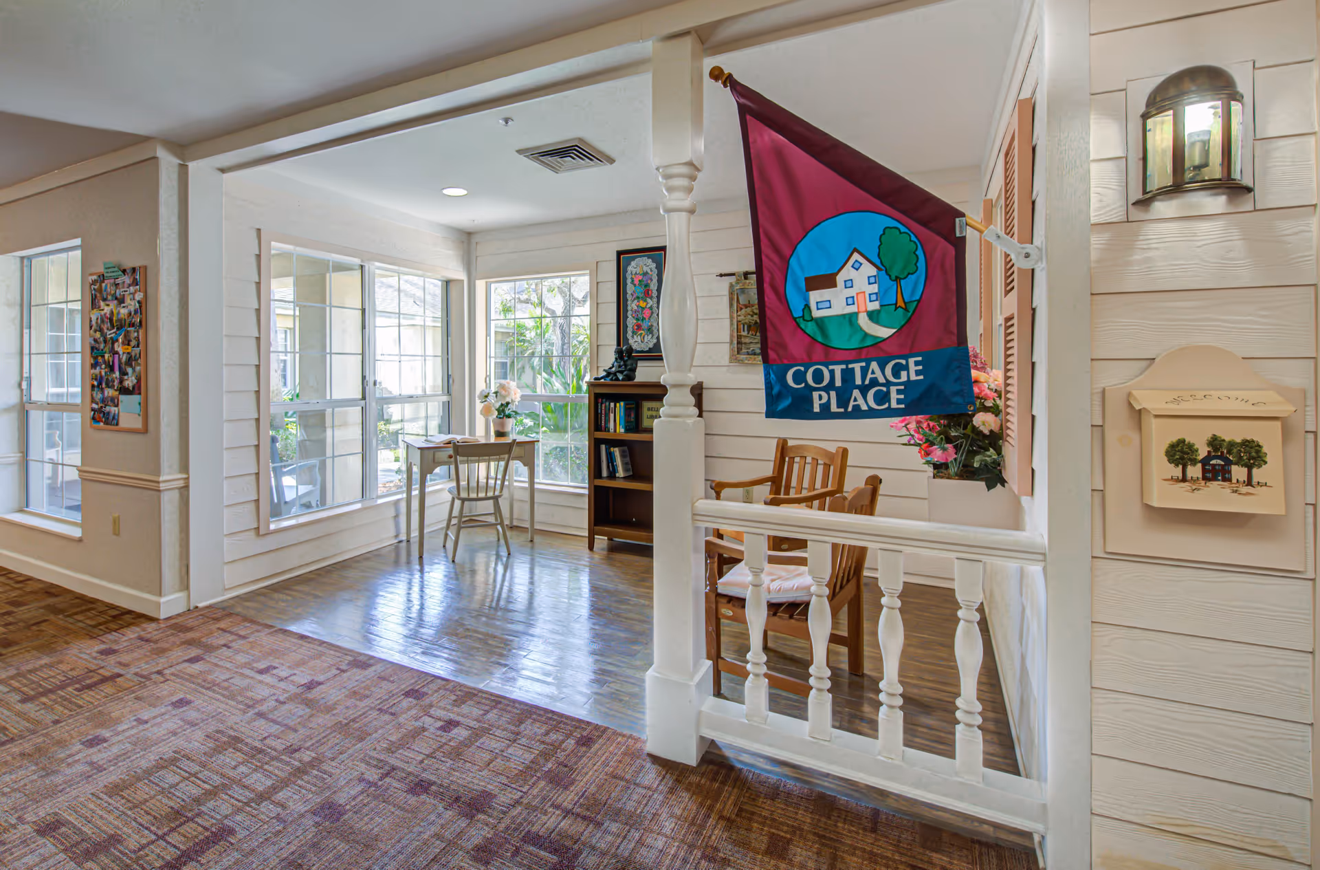 Interior view of a cozy sitting area in a senior living facility with large windows letting in natural light. The space features a small wooden desk with a chair, a bookshelf with books and decorations, two wooden chairs with cushions, and a colorful flag that reads 'Cottage Place' hanging on a white wooden railing. The walls are light-colored with decorative artwork and a small mailbox labeled 'Welcome'.