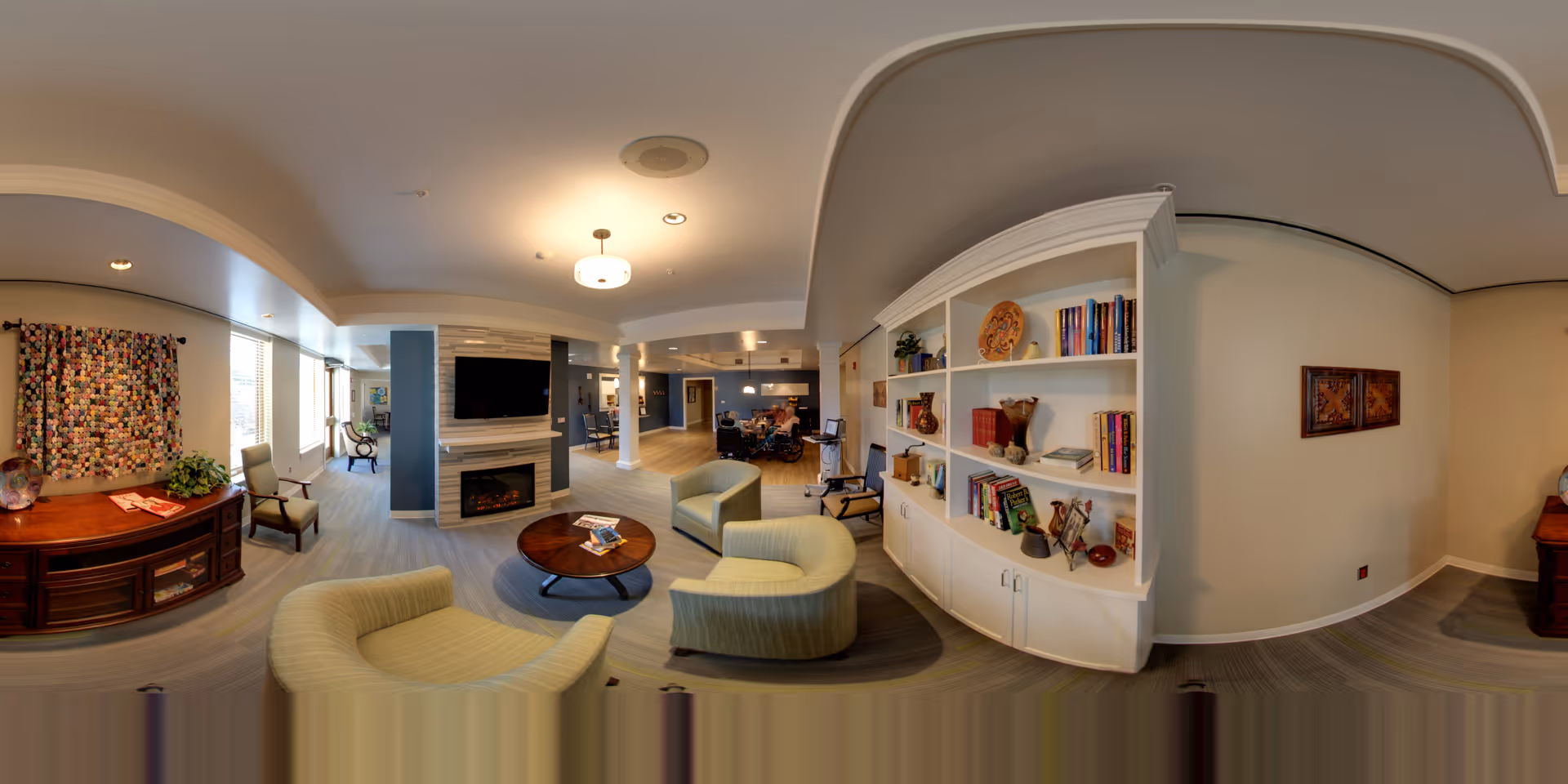 A spacious and well-lit living room area in a senior living facility with comfortable armchairs arranged around a round wooden coffee table. There is a white built-in bookshelf filled with books and decorative items on the right wall. A flat-screen TV is mounted above a fireplace in the center, and a wooden sideboard with a colorful quilt hanging above it is on the left. The room has a modern design with neutral tones and ample natural light coming through the windows.