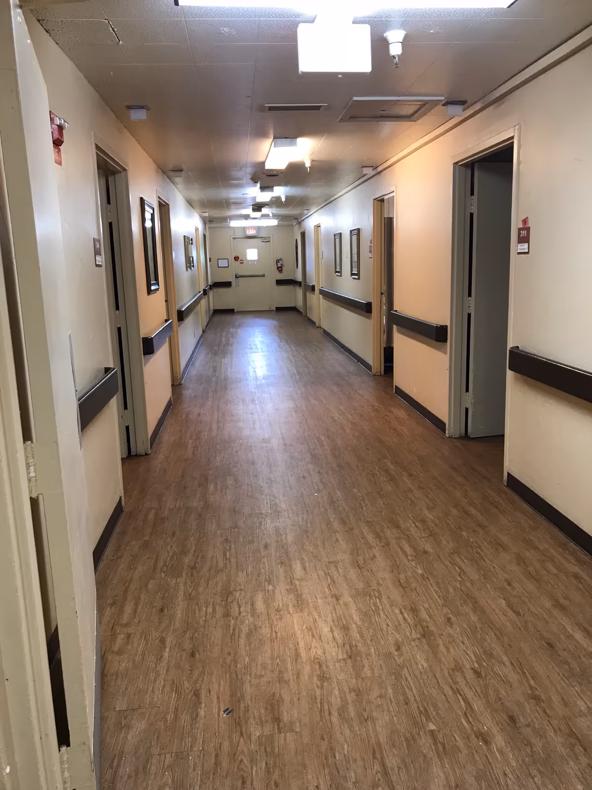 Long interior hallway with wood-look flooring, handrails and doors on both sides in a care facility.