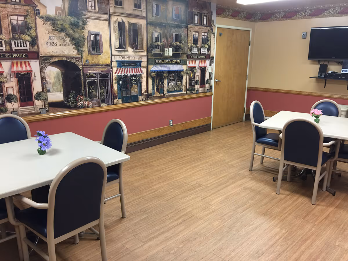 Interior of a dining area with two tables, each surrounded by four chairs with blue cushions. Each table has a small vase with flowers. The walls are painted beige and red with a mural depicting a European street scene with shops and buildings. A wooden door and a wall-mounted TV with a shelf below it are visible.