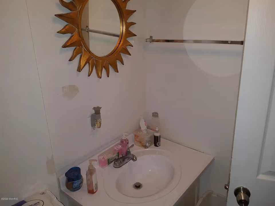 Small bathroom sink and countertop with a sunburst mirror, faucet, soap dispensers and toiletries.