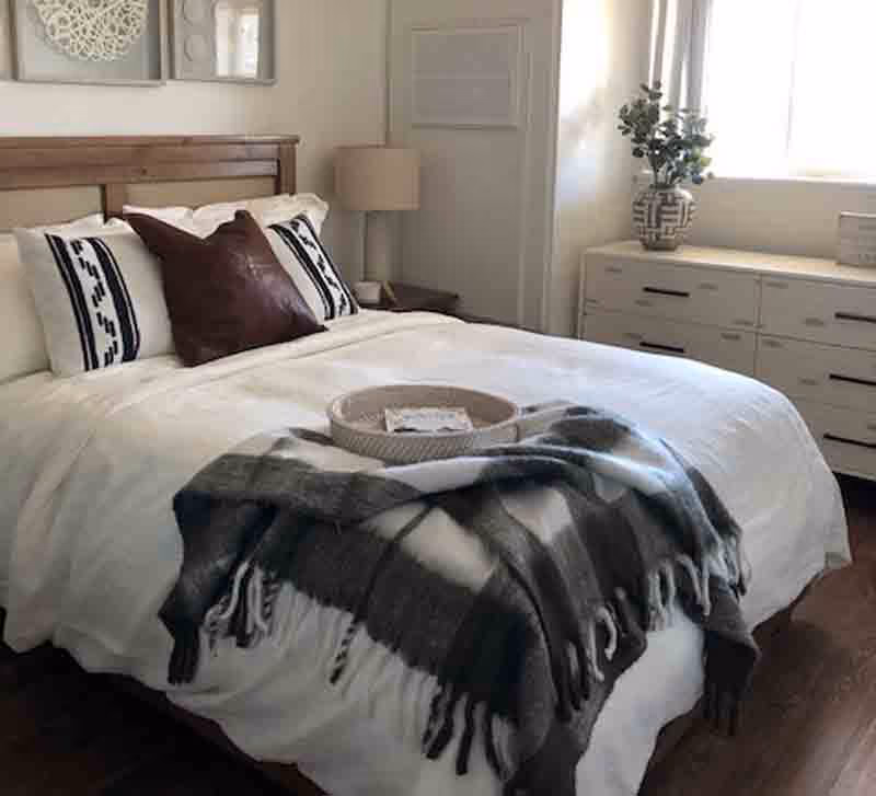 A neatly made bed with white bedding, two white pillows with black patterns, and a dark brown decorative pillow in the center. A gray and white plaid throw blanket is draped over the foot of the bed with a round woven tray on top. To the right, there is a white dresser with black handles, a decorative vase with greenery, and a window letting in natural light. A bedside table with a lamp is visible on the left side of the bed.