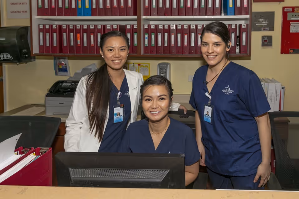 Three healthcare workers smiling behind a reception desk in an office setting. Two women are standing, one wearing a white coat and the other in navy blue scrubs, while a third woman in navy blue scrubs is seated in front of a computer monitor. Behind them are shelves filled with red binders and office equipment.