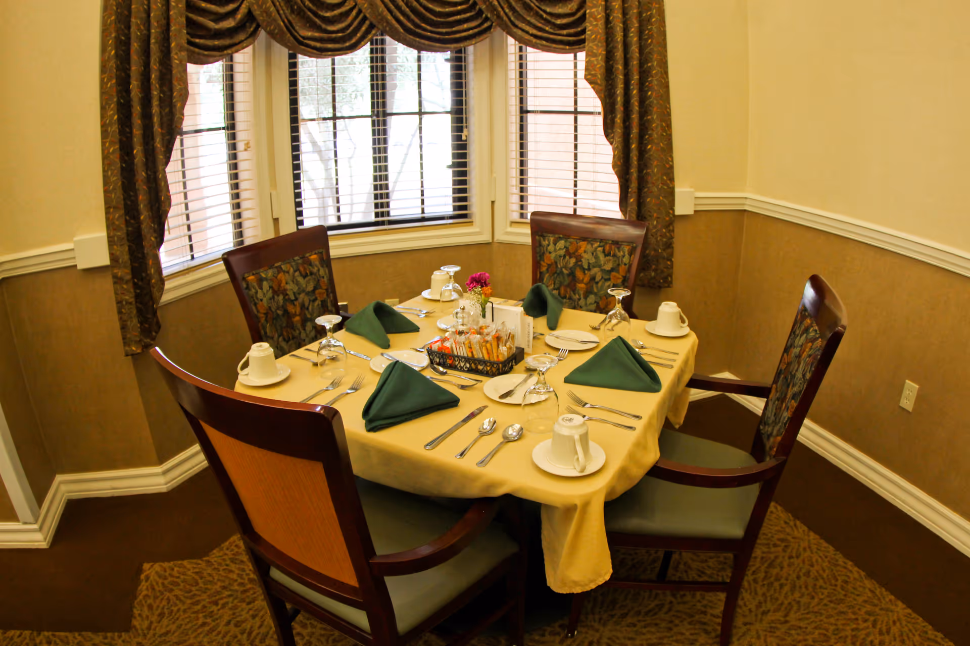 A dining table set for four people with a yellow tablecloth, green folded napkins, white cups and saucers, silverware, and a centerpiece with flowers and condiments. The table is surrounded by four wooden chairs with patterned upholstery. Behind the table are windows with blinds and brown curtains.