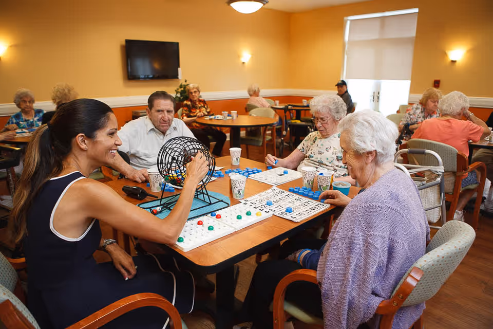 A group of elderly people and a caregiver sitting around tables in a well-lit room playing bingo. The caregiver is smiling and operating a bingo cage while the seniors focus on their bingo cards. The room has warm yellow walls, a wall-mounted TV, and several other seniors seated at tables in the background.
