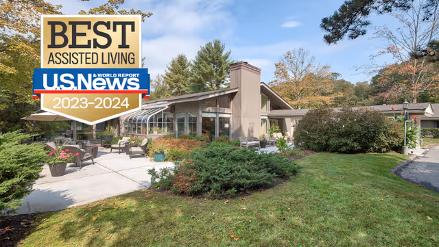 Outdoor view of a senior living facility with a patio area featuring chairs and tables surrounded by greenery and trees under a clear sky. The building has large windows and a sloped roof, with a well-maintained lawn and shrubs in the foreground.