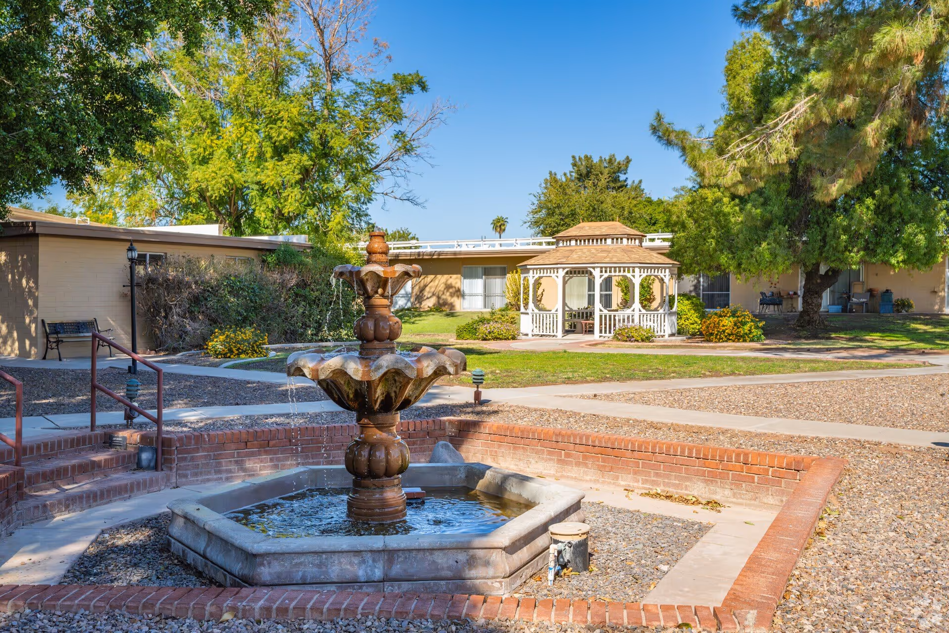 Outdoor area of a senior living facility featuring a multi-tiered water fountain in the foreground, surrounded by a brick border. In the background, there is a white gazebo with seating inside, green grass, trees, and shrubs under a clear blue sky.