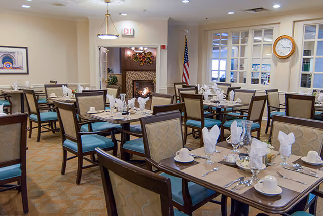 Dining room with multiple tables set with white napkins, cups, glasses, and silverware. Chairs with beige patterned backs and blue seats surround the tables. There is a fireplace in the background, an American flag, a clock on the wall, and framed artwork.
