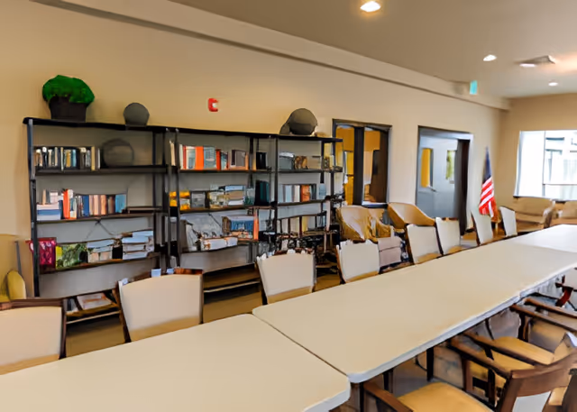 A large room with a long table surrounded by chairs. In the background, there are bookshelves filled with books and some decorative items. The room has beige walls, a window letting in natural light, and an American flag near the window.