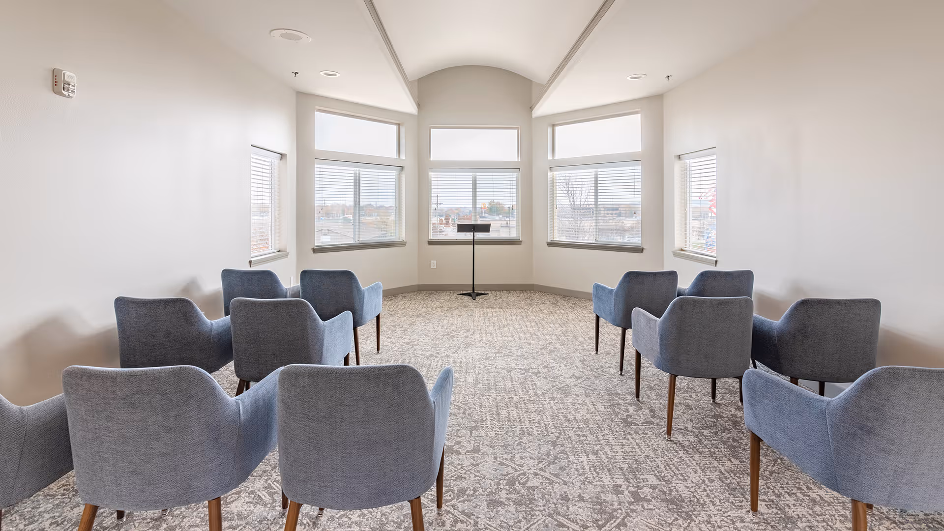 A small meeting or presentation room with nine blue upholstered chairs arranged in three rows facing a music stand near a set of large windows with blinds. The room has light-colored walls and a patterned carpet floor.