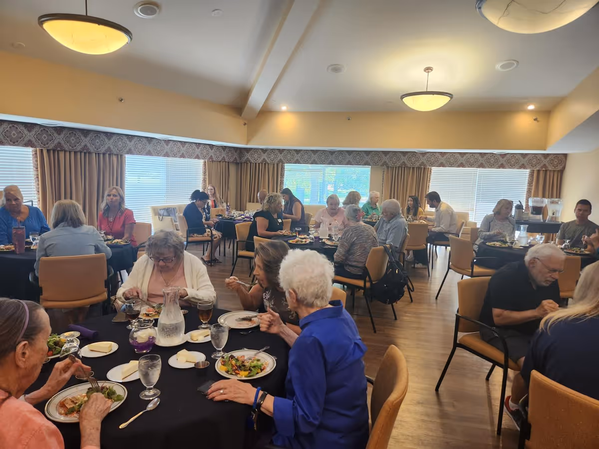 A group of elderly people and some staff members sitting at round tables in a dining room, eating meals and socializing. The room has large windows with curtains, warm lighting, and wooden flooring.
