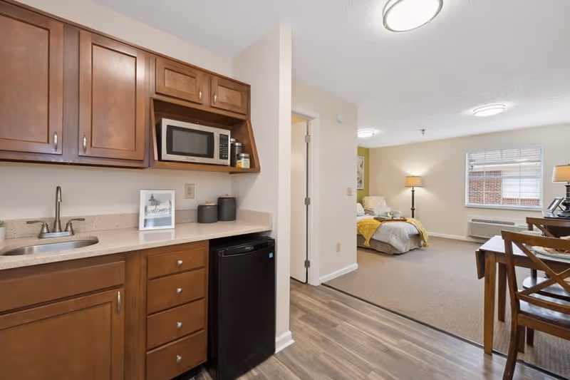 Interior view of a senior living apartment showing a kitchenette with wooden cabinets, a microwave, a small refrigerator, and a sink. The kitchenette opens into a living and sleeping area with a bed, a chair, a floor lamp, a window with blinds, and a dining table with chairs.