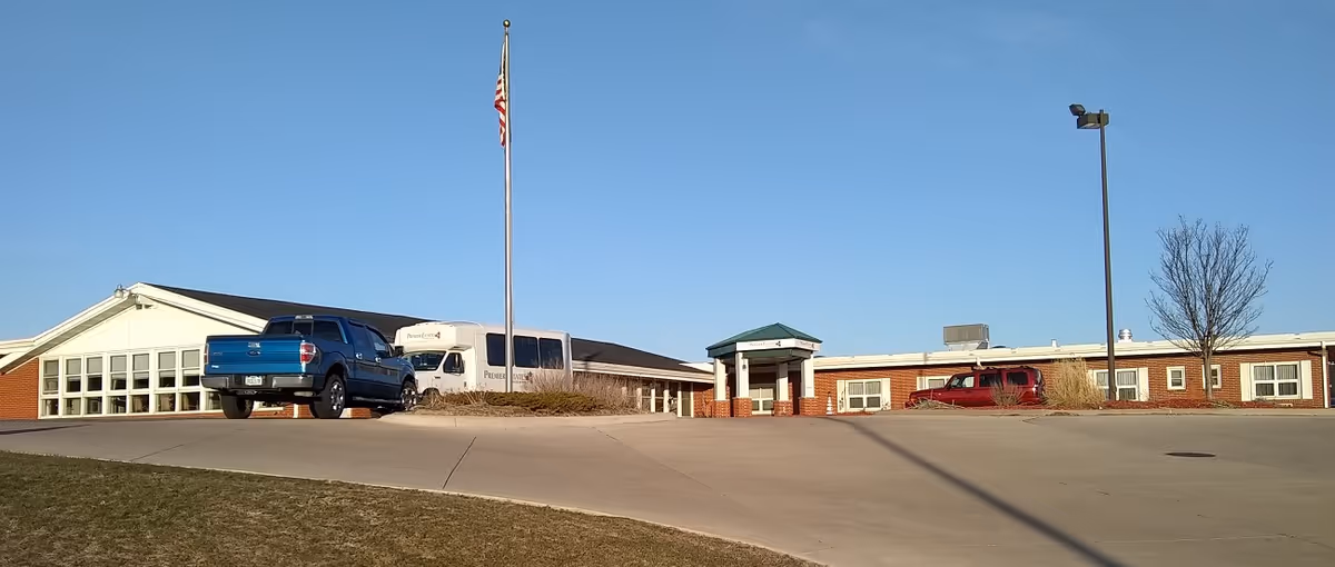 Single-story brick building front with an entrance canopy, flagpole, and parked vehicles under a clear blue sky.