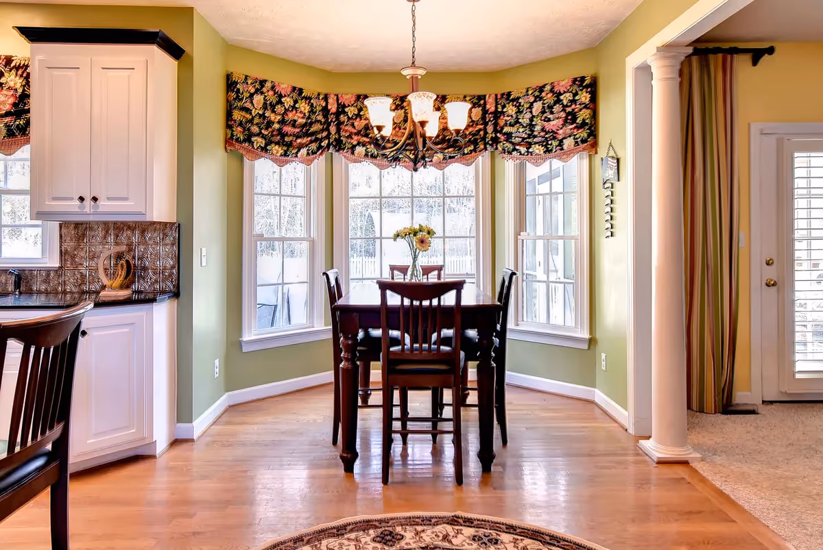 A dining area with a wooden table and four chairs placed in front of large bay windows with floral valances. A chandelier with five lights hangs above the table, which has a vase with flowers on it. To the left, part of a kitchen counter and cabinets are visible, and to the right, a column separates the dining area from a carpeted living space with a door and striped curtains.