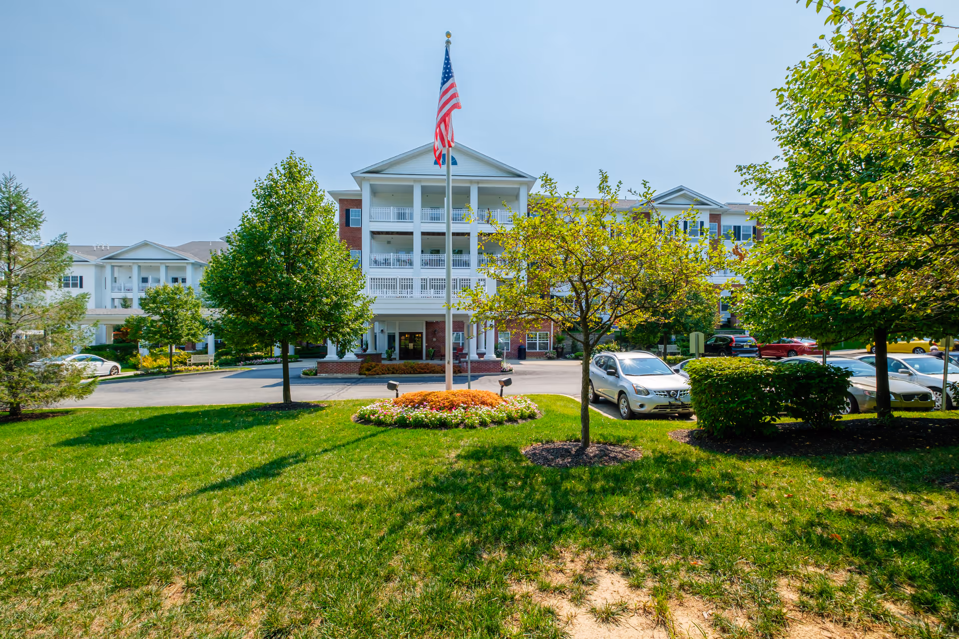 Front exterior view of a senior living facility with a large American flag on a flagpole in the center, surrounded by green grass, trees, and parked cars. The building has white balconies and brick accents under a clear blue sky.