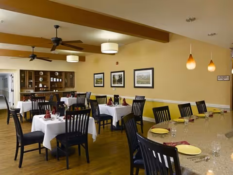 Dining room with several tables covered in white tablecloths, set with plates, glasses, and folded napkins. Black wooden chairs surround the tables. There is a counter with yellow plates and glasses on the right side. The room has wooden floors, beige walls with three framed pictures, ceiling fans, and hanging pendant lights.