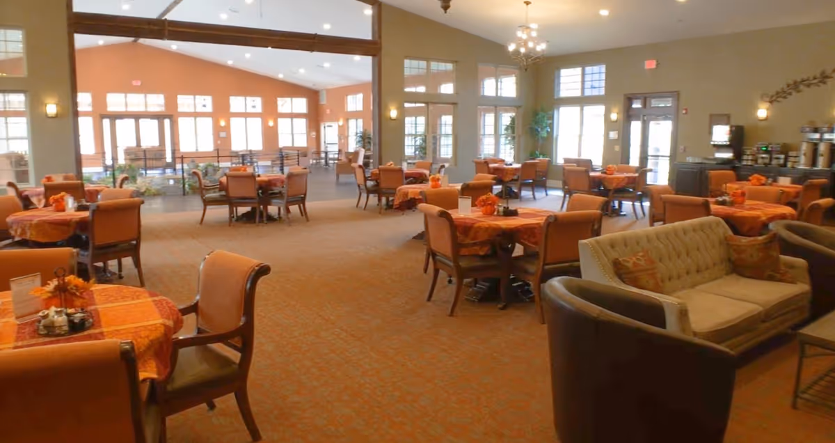 A spacious dining area with multiple tables covered in orange and red tablecloths, each surrounded by cushioned chairs. The room has large windows allowing natural light to fill the space. There is a seating area with a beige tufted sofa and dark armchairs in the foreground. The walls are painted in warm tones, and there are plants and a beverage station visible in the background.