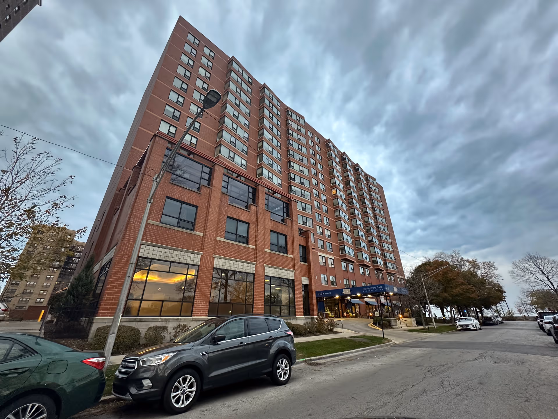 Exterior view of a multi-story brick building under a cloudy sky with several parked cars along the street. The building has large windows on the lower floors and a blue awning above the entrance that reads 'Montgomery Place Retirement Community.' Trees line the street on the right side.