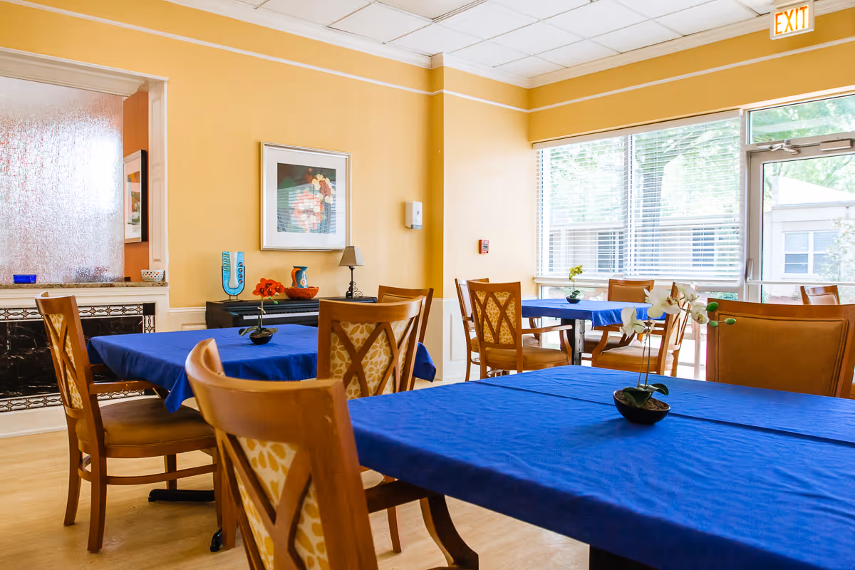 A bright dining room with several wooden tables covered with blue tablecloths and matching wooden chairs. Small potted plants are placed on the tables. The room has yellow walls, a large window with blinds letting in natural light, and an exit door. There is a framed picture on the wall and a small side table with decorative items.