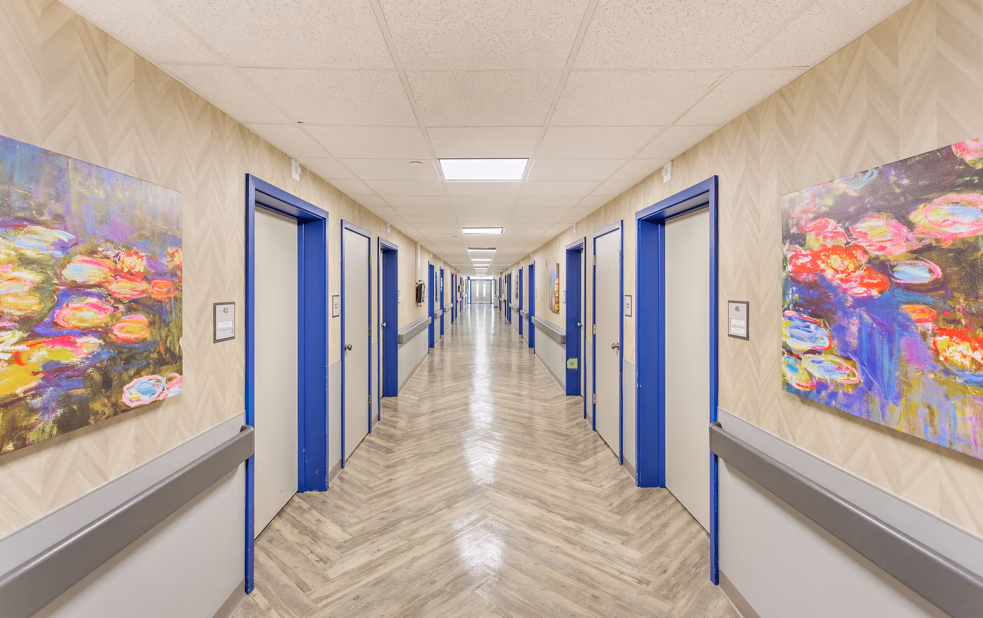A long, well-lit hallway in a senior living facility with multiple closed doors on both sides, each door framed in blue. The walls have a beige patterned wallpaper with colorful abstract paintings hanging on either side. The floor has a wood-like herringbone pattern, and there are handrails along the walls.