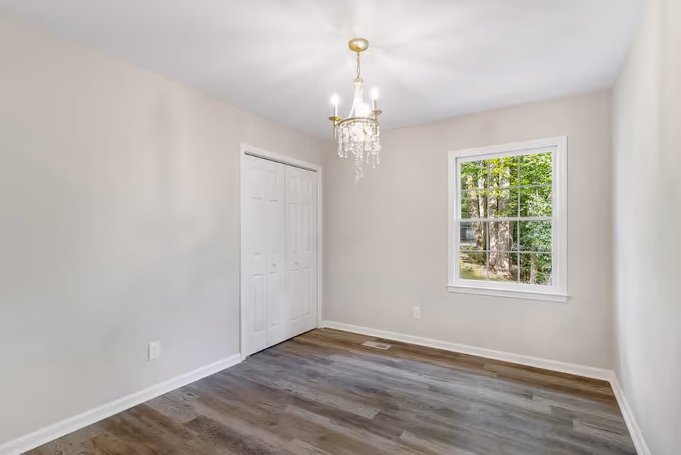 Empty room with light gray walls, a window showing green trees outside, a white double-door closet, wood flooring, and a chandelier hanging from the ceiling.
