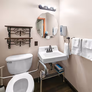 Small accessible bathroom with a toilet and grab bars, wall-mounted sink under a round mirror, towel rack, decorative shelving and soap dispensers.