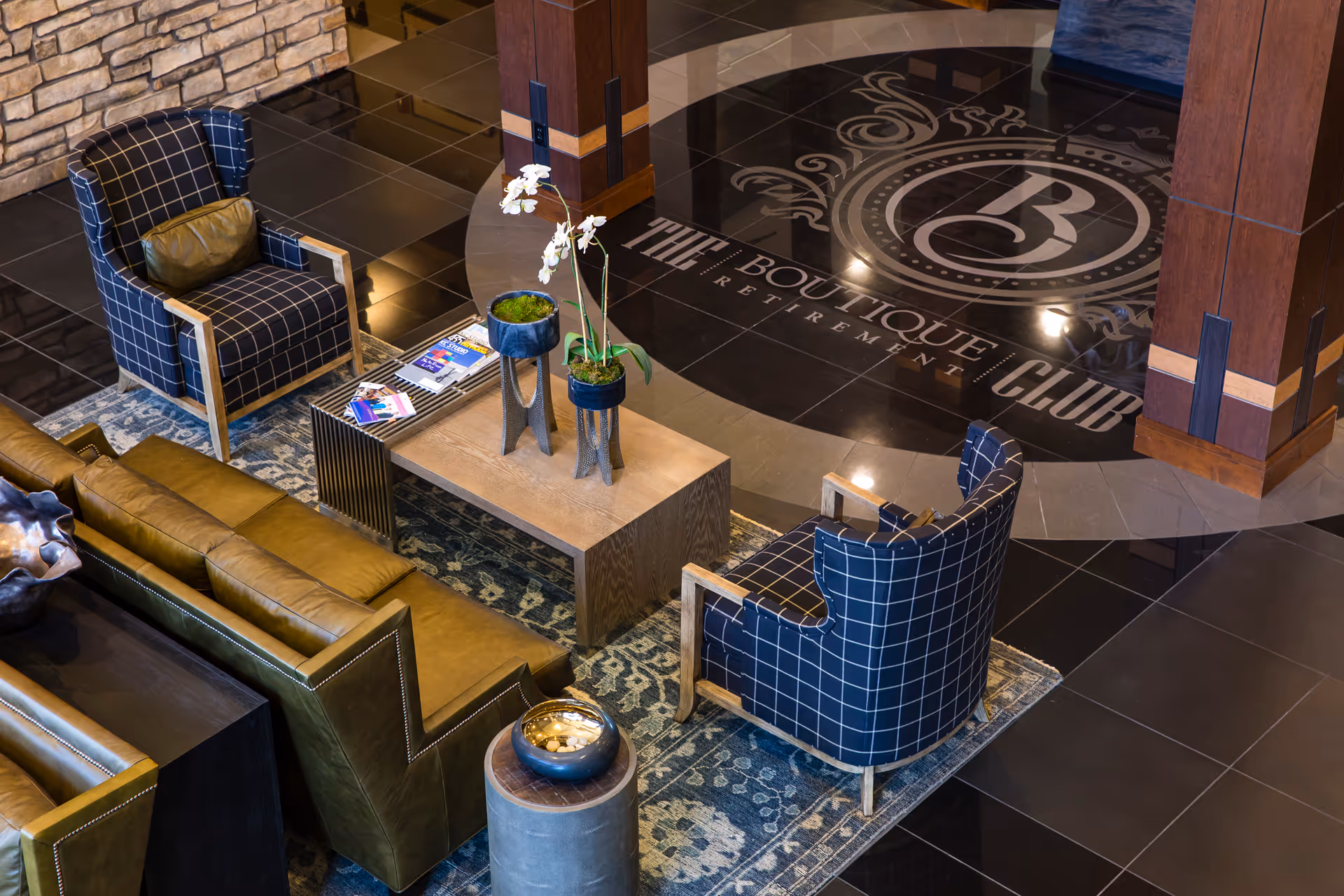 A stylish seating area with two blue plaid armchairs and a green leather sofa arranged around a wooden coffee table with potted plants on top. The floor features a decorative circular tile design with the text 'The Boutique Club Retirement'.