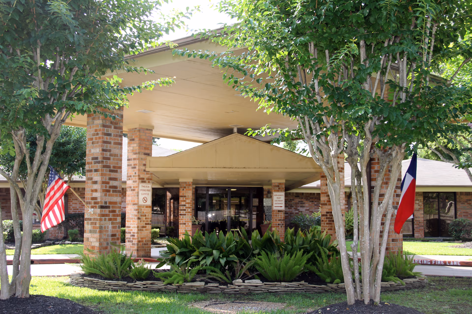 Entrance of Baywind Village Skilled Nursing and Rehabilitation facility with a covered drop-off area supported by brick pillars, surrounded by green trees and plants. American and Texas flags are displayed on either side of the entrance.