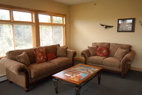 A cozy living room with two patterned brown sofas facing each other, adorned with decorative pillows. A rectangular coffee table with a colorful puzzle or artwork on top is placed between the sofas. Large windows with wooden frames and blinds allow natural light to fill the room. The walls are light-colored with minimal decoration, including a small framed picture and a wall-mounted decorative piece.
