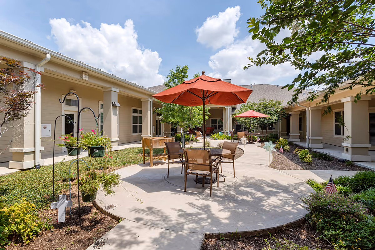 Sunny courtyard with tables and red umbrellas surrounded by a single-story assisted living building and landscaped walkways.
