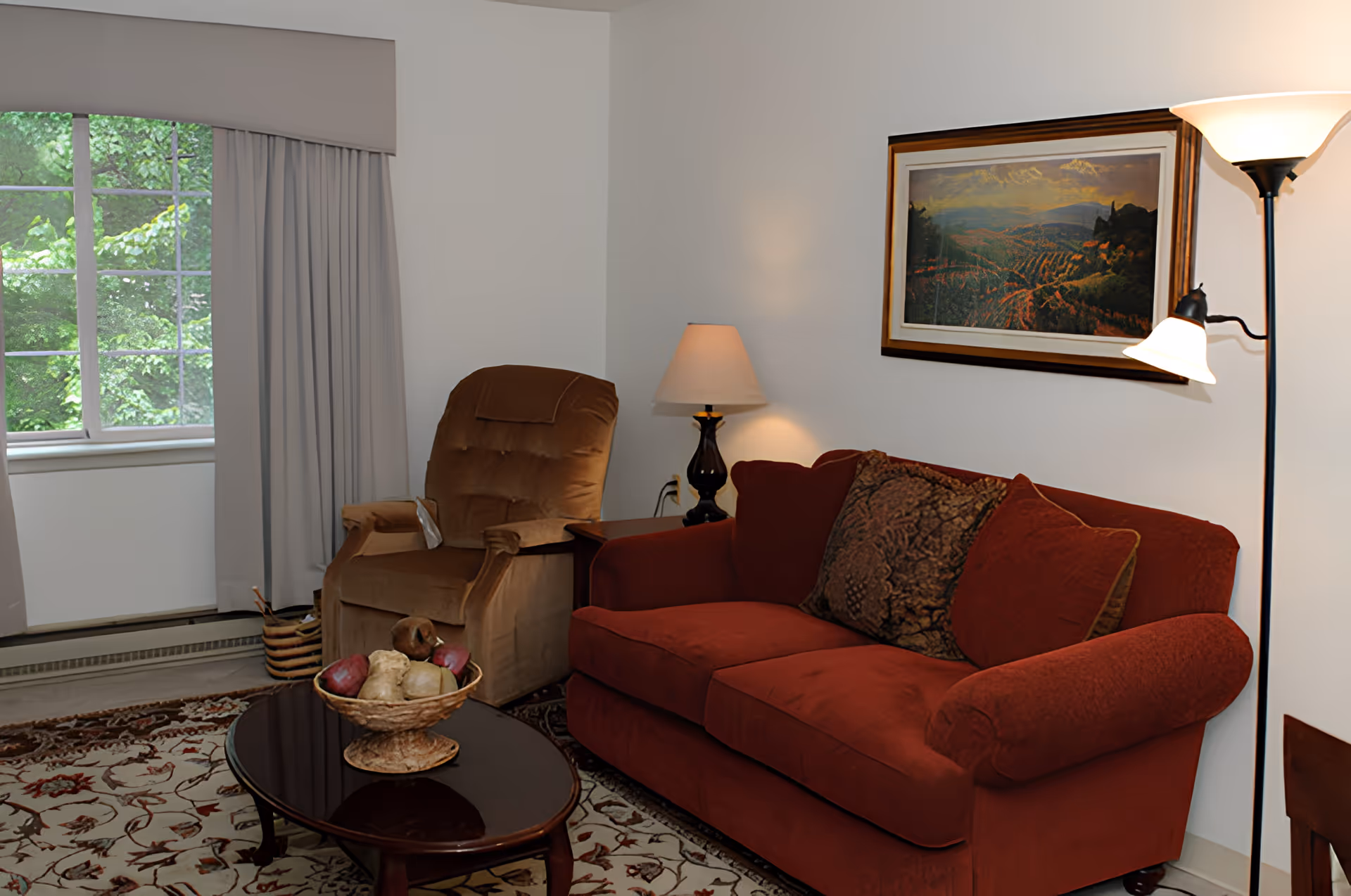 A cozy living room with a red sofa adorned with patterned cushions, a brown recliner chair, a wooden coffee table with a decorative bowl of artificial fruit, a floor lamp, a table lamp on a side table, a framed landscape painting on the wall, and a window with gray curtains showing green trees outside.