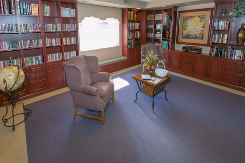 A cozy library room with built-in wooden bookshelves filled with books, two upholstered armchairs, a small wooden coffee table with a flower arrangement and a basket, a globe on a stand, and a window with blinds letting in natural light.