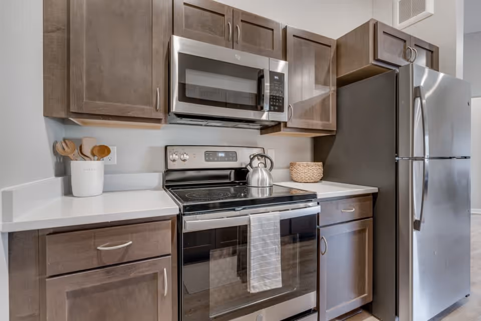 Modern kitchen area with stainless steel appliances including a microwave, stove with a kettle on it, and a refrigerator. The kitchen has brown wooden cabinets and white countertops with a utensil holder and a woven basket on the counter.