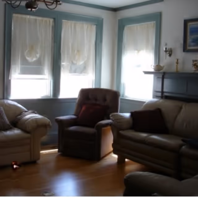 A cozy living room with three upholstered chairs and sofas arranged around a wooden floor. Three windows with sheer white curtains allow natural light to enter the room. The walls are light-colored with blue trim, and there is a dark-colored mantel with decorative items on it.