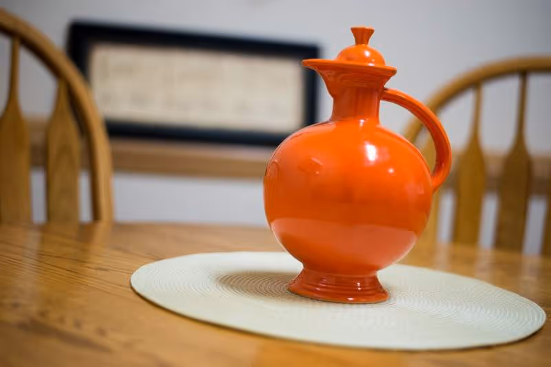 A bright orange ceramic pitcher with a handle and lid sits on a round white placemat on a wooden dining table. In the background, there are wooden chairs and a framed picture on the wall.