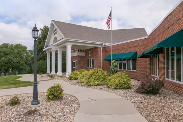 Exterior view of Colonial Manor Of Randolph, a brick building with white columns at the entrance, an American flag on a flagpole, green awnings over windows, a curved concrete walkway, landscaping with bushes and rocks, and a black lamp post.