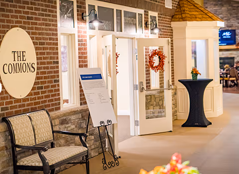 Interior common area showing a brick-wall sign reading 'The Commons,' a bench, open double doors with wreaths, a standing display sign, and a cocktail table with flowers.