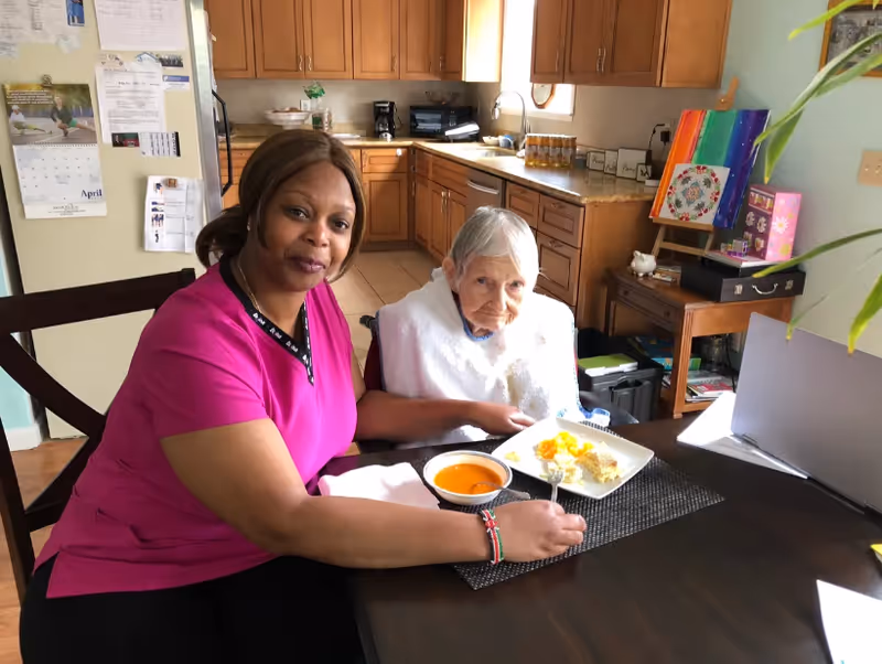 A caregiver in a pink uniform sitting at a dining table with an elderly woman who is wearing a white bib. The elderly woman has a plate of food and a bowl of soup in front of her. The background shows a kitchen area with wooden cabinets and a small table with colorful artwork and various items.