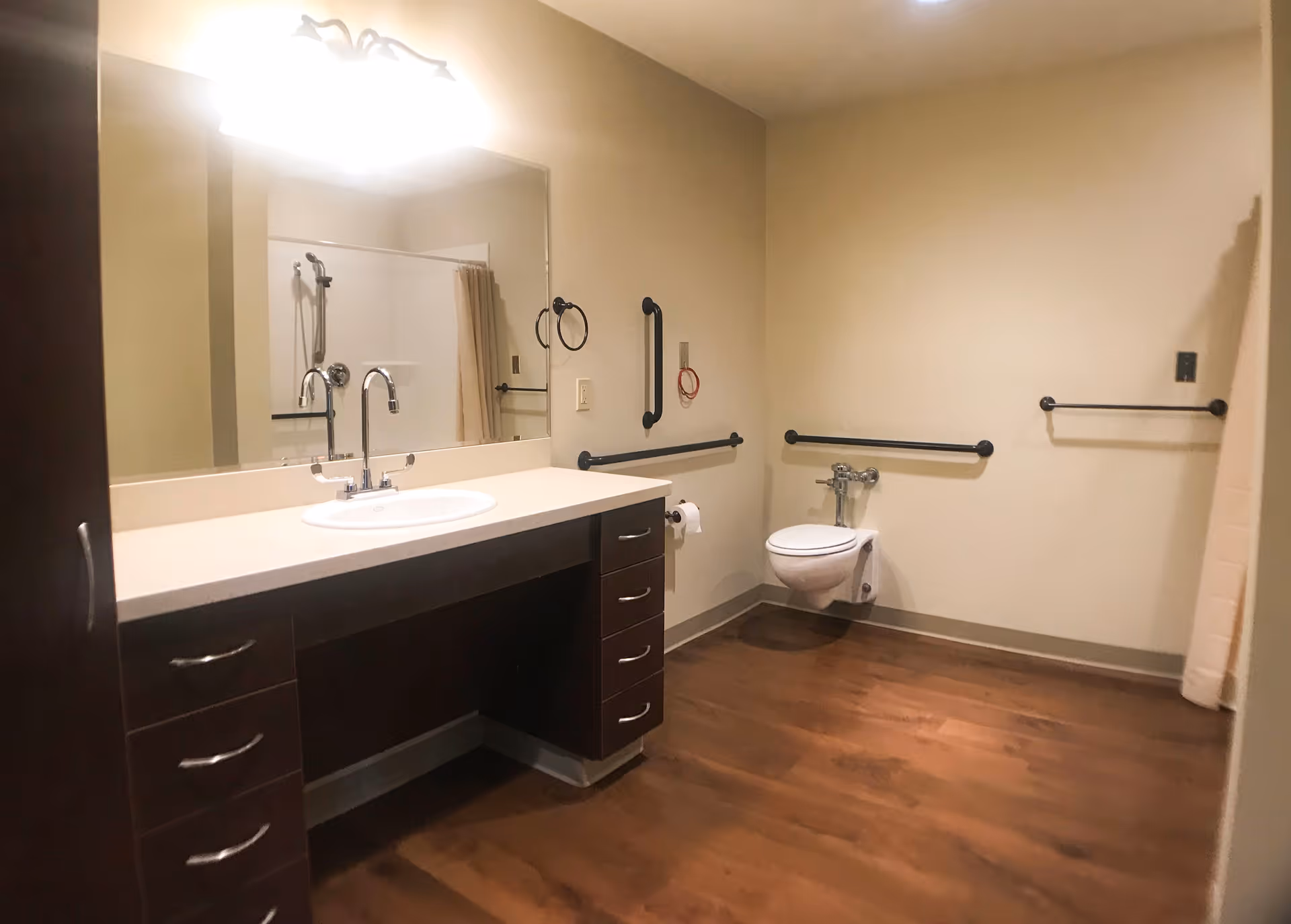 A spacious bathroom with a large mirror above a sink with a countertop and dark wood cabinetry. The room features a wall-mounted toilet with grab bars on the walls for accessibility, a shower area with a curtain, and wood-look flooring.