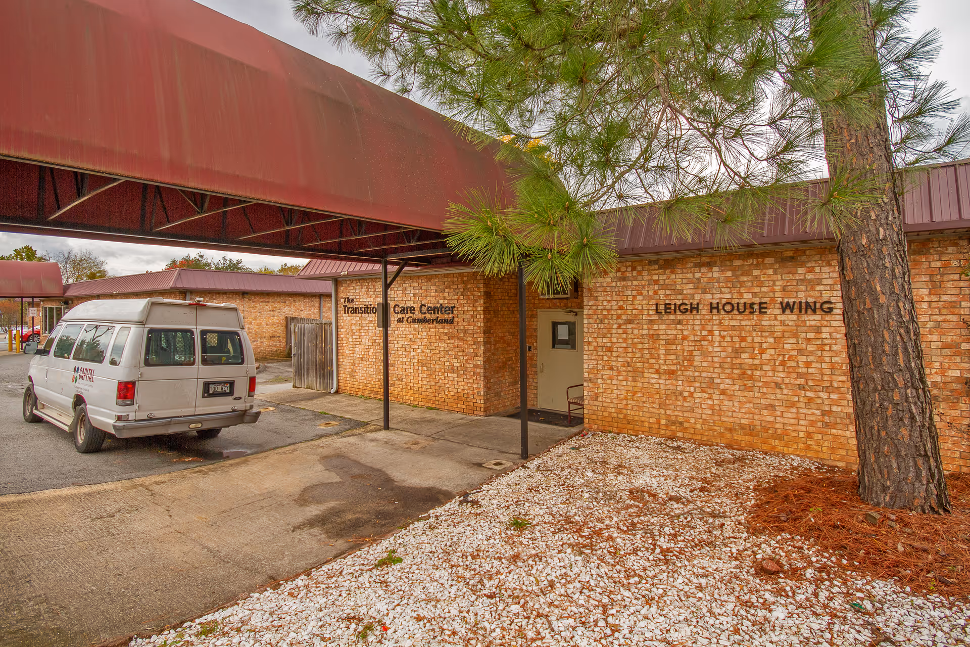 Exterior view of a brick building with a covered entrance. The building has signs reading 'The Transition Care Center at Cumberland' and 'Leigh House Wing'. A white van is parked under the covered area, and a tree with green needles is visible on the right side near the building.