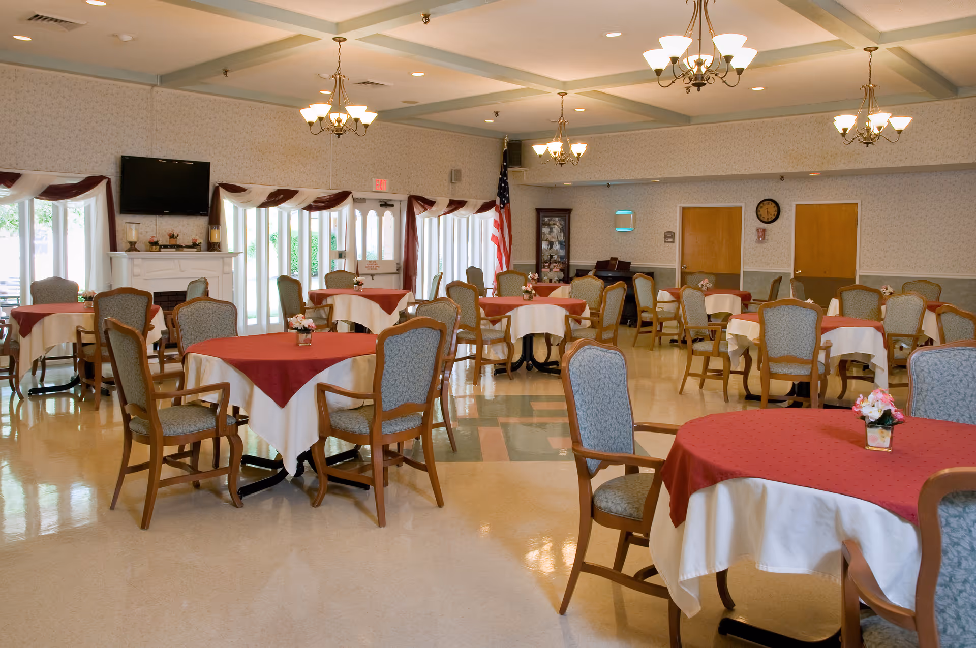 A spacious dining room with multiple round tables covered with white tablecloths and red overlays. Each table is surrounded by wooden chairs with cushioned seats and backs. The room features chandeliers hanging from the ceiling, large windows with curtains, an American flag, a wall-mounted TV above a fireplace, and a clock on the far wall.