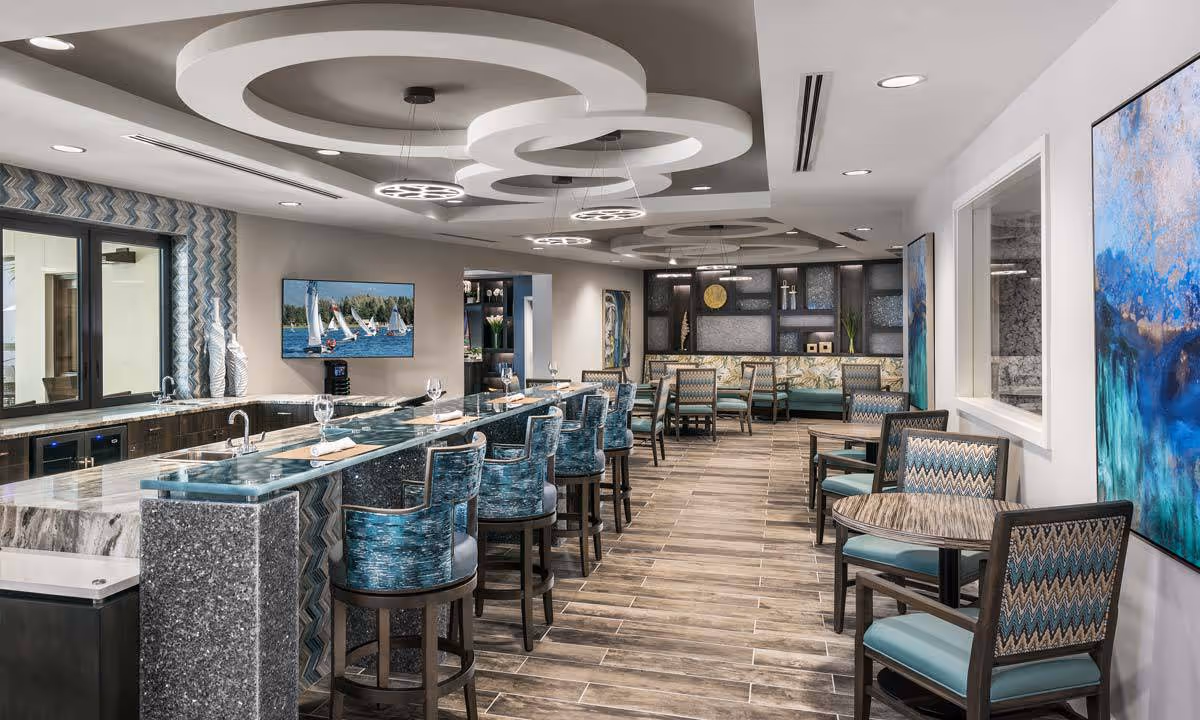 A modern dining area with a long marble and granite bar counter lined with blue cushioned bar stools. The room features multiple round tables with chairs upholstered in blue and patterned fabric. The ceiling has circular recessed lighting fixtures, and the walls are decorated with colorful abstract paintings. A flat-screen TV displaying sailboats on the water is mounted on one wall.